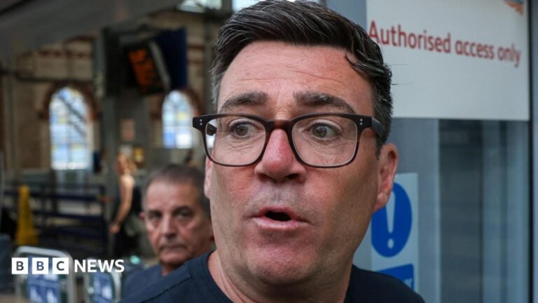 Andy Burnham rules out reform of Wales funding system Andy Burnham pictured in the centre of the frame, wearing dark-framed glasses and a dark navy or black top. The photograph is tightly cropped around the head and shoulders. His face is sharply in focus, while most of the background is blurred. Behind him is a railway station concourse. A second man is visible in the background, but out of focus.