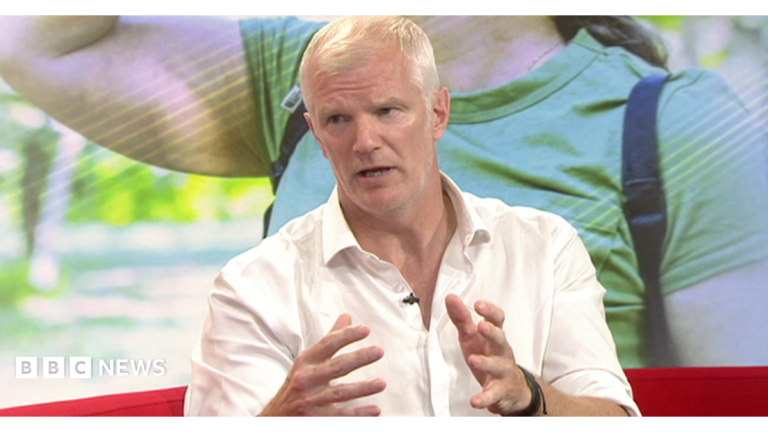 A man with short white hair is wearing an open-neck white shirt and sitting on a red sofa in a TV studio. There is a photographic backdrop behind him illustrating the heatwave, with a woman in summer clothes and a backpack.