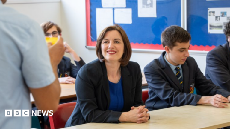 Phillipson wears a black suit jacket over a royal blue dress with a silver necklace. Her hair is worn in a dark brown, chin-length bob. She wears bright red lipstick and is smiling at a teacher, who can partially be seen in the foreground on the left. There are pupils sitting around her, all wearing a navy school uniform with a pastel blue shirt and a navy tie with pale blue and gold diagonal stripes.