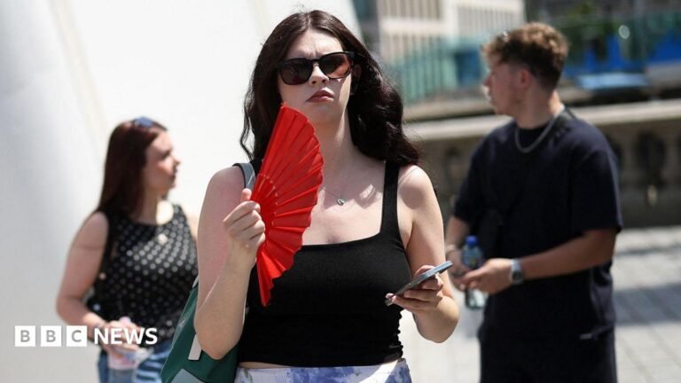 Hundreds of schools plan closures ahead of red heat alerts A woman on the street waves a red fan and holds her phone. She wears sunglasses and a black top.