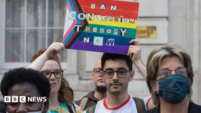 Jail time and unlimited fines planned under conversion practices ban A tightly cropped image of campaigners protesting for a ban on conversion practices. Two campaigners are in sharp focus, holding a sign featuring the progress pride flag and the words 'Ban Conversion Therapy Now' written in letters cut out from a newspaper or magazine. Both protestors are wearing glasses and have serious looks on their faces. Other protestors in the foreground are slightly out of focus.
