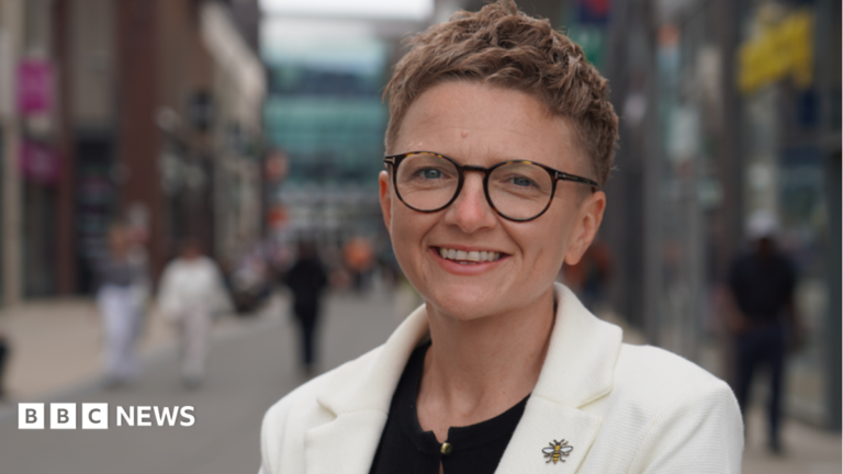 Bev Craig has short red hair, and is wearing glasses and a white jacket over a black blouse