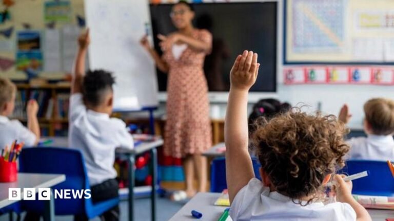 Number of children getting special educational needs support hits record high Children in a classroom sit at desks with hands raised while a teacher stands at the front beside a whiteboard, leading a lesson. Colourful posters decorate the walls and stationery items are visible on desks.