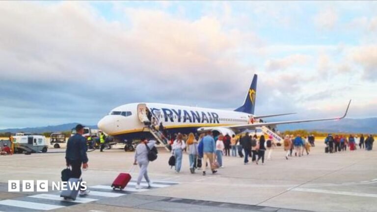 Passengers line up to board a Ryanair aircraft on an airport runway under a colourful sunset sky.