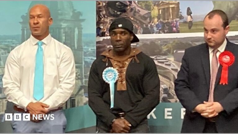 Three men at a local election for Wakefield in May 2026 - a tall, bald man is on the left wearing a white shift, turquoise tie and blue trousers; the man in the centre has a black baseball cap on backwards and is wearing a black and brown hoody with black jeans; the man on the right has short black hair, a black suit and a pink tie. All three men have their hands clasped in front of them.