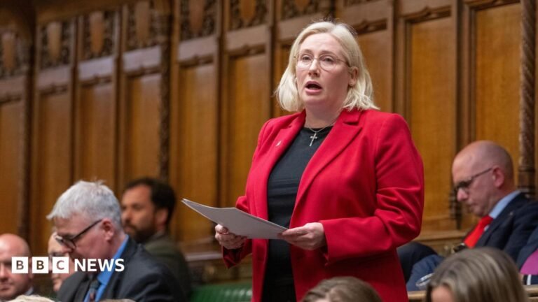 Emma Lewell MP stands giving a speech in front of the wood panelled walls of the House of Commons chamber surrounded by colleagues. She's wearing a crisp bright red blazer over a black top and has a silver crucifix necklace on. She's mid sentence holding papers in her hands in front of her.