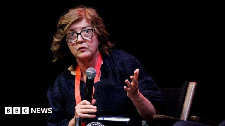 Baroness Sue Gray sitting on a chair holding a microphone. She wears a red lanyard and has short brown hair and glasses.