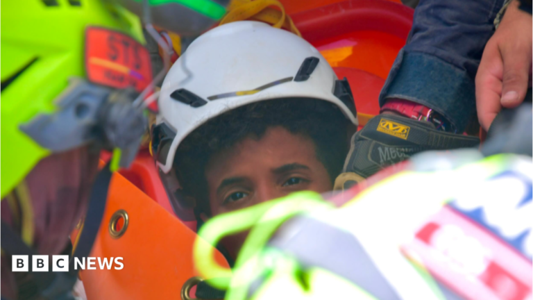the picture shows half of the boys face - the rest is obscured by rescue workers. He's wearing a white hard hat and has dark curly hair.