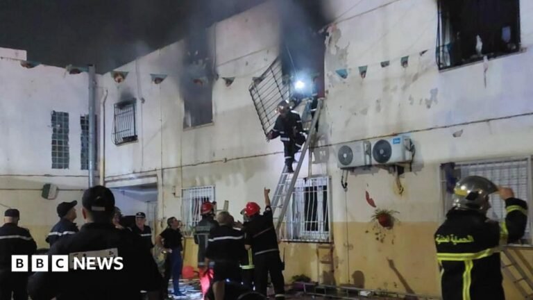 A fireman up a ladder removing a grill from a window from which black smoke is billowing. He is gesturing to other fire officials below who are pointing - some wearing red hard hats.