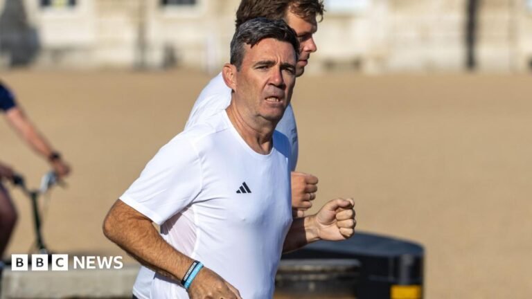 Andy Burnham wearing a white Adidas t shirt and black athletic shorts, running next to Green Park. The sun is on his face