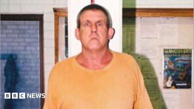 A tall man in a yellow t-shirt and blue short stands against a measuring tape in an office