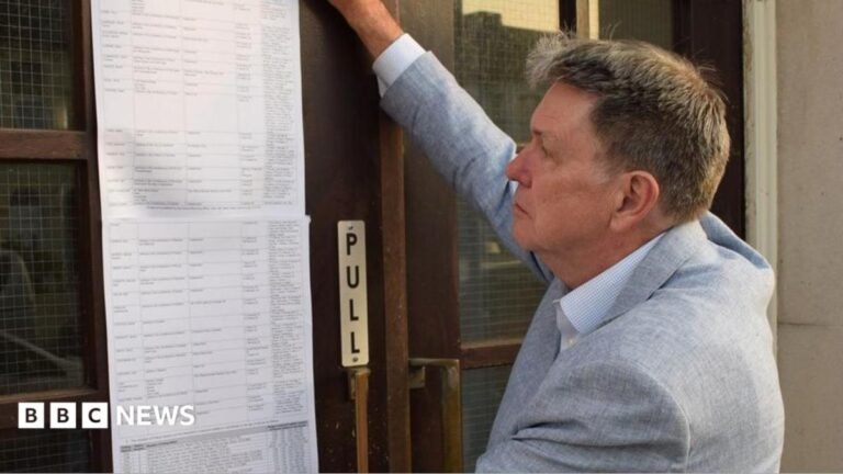 Clacton by-election attracts 34 candidates Ian Davidson posting the statement of persons nominated outside Clacton Town Hall. He is on the right, has short light brown hair and is wearing a light grey jacket over an open-neck shirt. His right arm is upraised as he puts the long list of typed names on to the brown wooden and glass door.