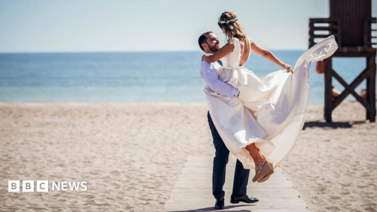 Couples free to marry at the beach or the pub under new plans A man lifts up a woman dressed in a wedding dress on a boardwalk on a beach. In the background is the ocean and a lifeguard chair.