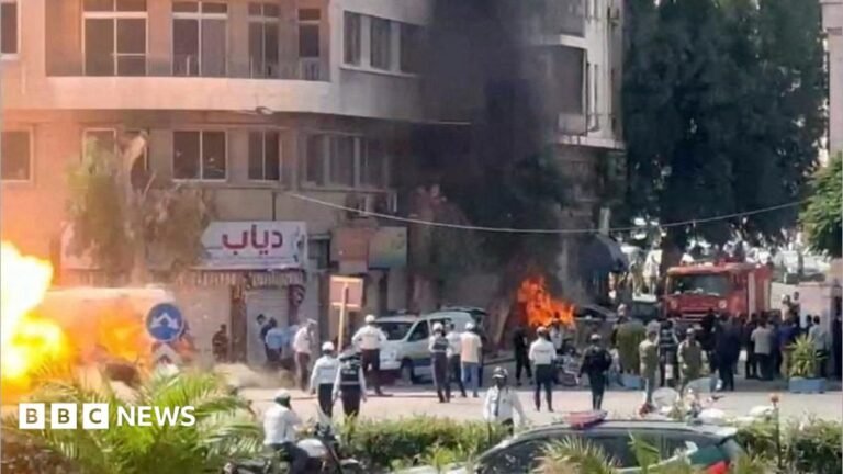 Explosions injure 18 in Damascus during Macron’s visit A wide shot of a street with lots of emergency workers. Flames and smoke rise from an alleged explosion site