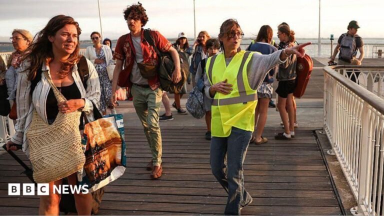 A volunteer gives indication to holidaymakers from Lege-Cap-Ferret upon their arrival in Arcachon by boat during an evacuation operation of the entire Cap-Ferret peninsula. The tourists are seen panicked on a wooden getty while the holidaymaker is in a hi-vis jacket. The sea is seen in the background.