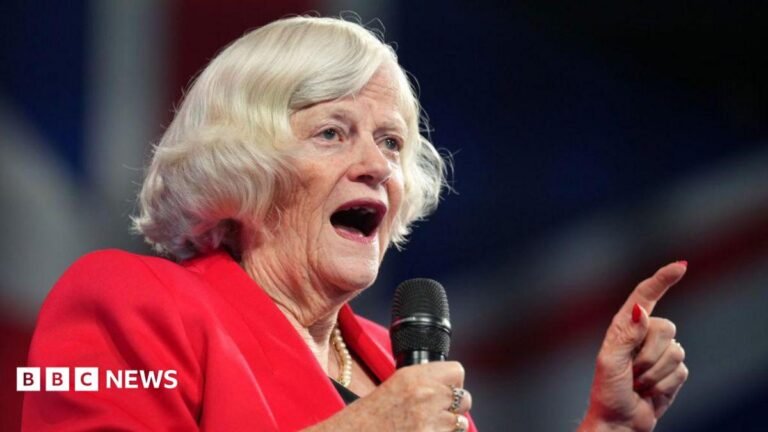 A picture of Widdecombe and Du Beke dancing in the Strictly Come Dancing studio. Anton Du Beke wearing a black shirt and trousers and a purple tie. He is spinning Ann Widdecombe around, holding her by the hand and foot. Widdecombe is wearing a glittery golden dress with purple sleeves.