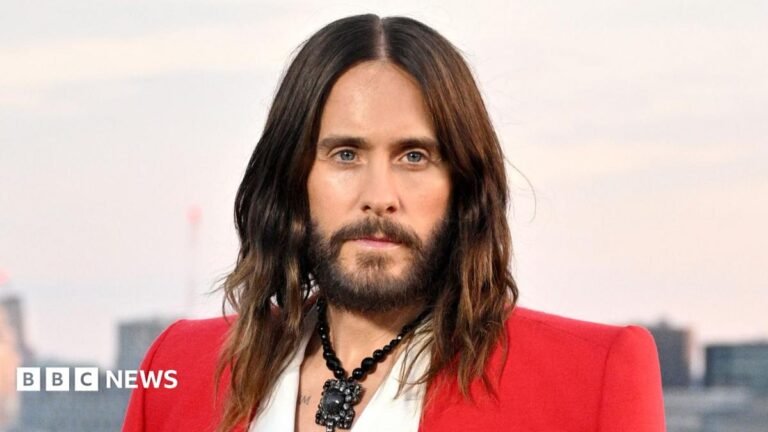 Four women accuse Jared Leto of criminal sexual conduct when they were teenagers A close-up portrait of Alex looking serious, with pursed lips. Her face is turned to the side and she is looking towards the right of the frame. She has blonde hair and wears a dark top and small earring. The background is softly blurred in shades of yellow, gold, and dark brown.