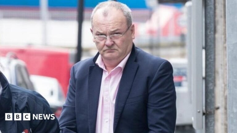 A man with thin greying hair and glasses in a pink shirt and dark blue suit