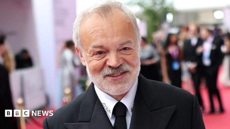 Graham Norton at the Baftas 2026 on the red carpet, wearing a black suit and tie and a white shirt with diamante detail on the collar