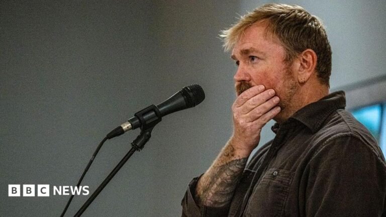 Graham Platner seen on stage at a microphone. his hand is over his mouth