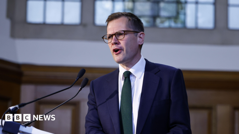 Jenrick urges Burnham to rule out tax rises to fund social care Reform UK's Robert Jenrick mid-speech. He is wearing glasses, has short, dark hair and is wearing a collared shirt and suit with a green tie.