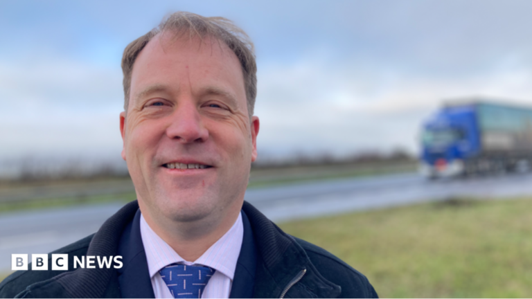 A man wearing a blue jacket and a blue tie stands in front of a road. He has receding light brown hair and is smiling at the camera. There is a HGV entering the picture in the background.