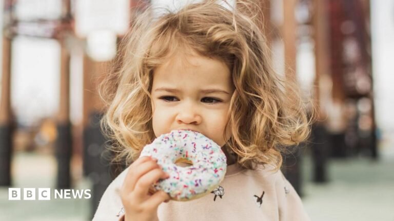 The image shows a young child outdoors holding a large doughnut with white icing and colourful sprinkles. The child is taking a bite from the doughnut, which occupies much of the foreground. The background contains blurred structural elements, such as columns or beams, creating a shallow depth-of-field effect. Soft natural lighting highlights the child's curly hair and the pastel tones of the clothing and doughnut icing.