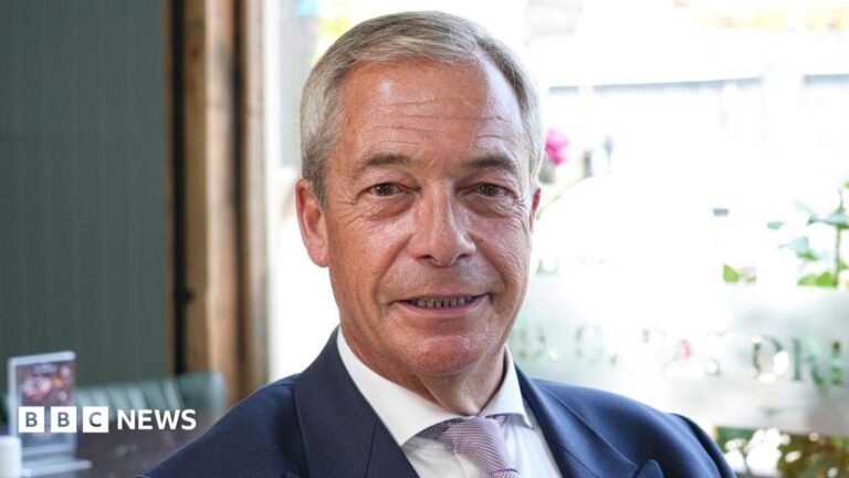 Nigel Farage sitting in a pub while wearing a white shirt and navy blazer. He is smiling.