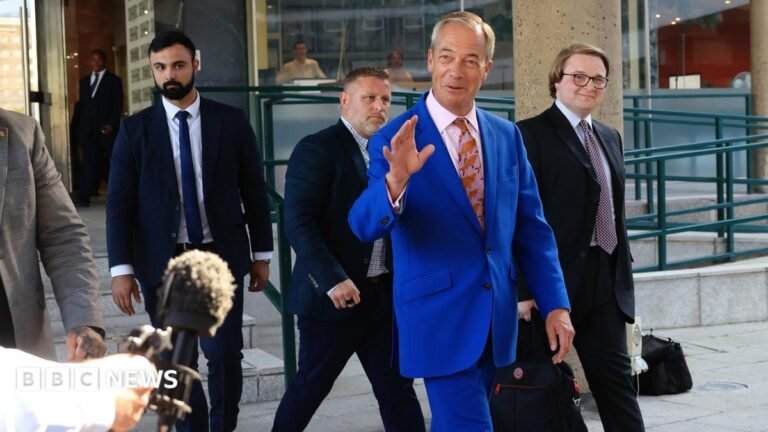 Nigel Farage walking outside, wearing a blue suit with a pink tie, decorated with a fish pattern. There are two men behind him, both wearing dark suits.
