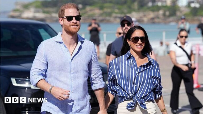 Prince Harry, Duke of Sussex, and his wife Meghan, the Duchess of Sussex walking hand in hand, wearing blue shirts and sunglasses.