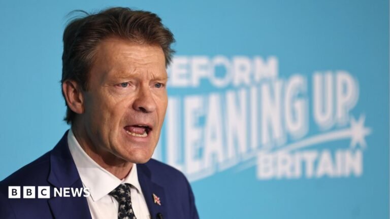 Richard Tice in a dark suit, wearing a union flag button on his lapel. He is standing behind a large turquoise blue sign with white writing which reads: Reform cleaning up Britain