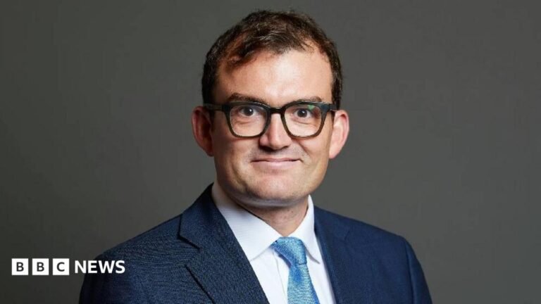 Suffolk MP Patrick Spencer not guilty of sexual assault Patrick Spencer wears a navy suit and stands in front of a plain background. He is smiling directly at the camera and is wearing large black-rimmed glasses.