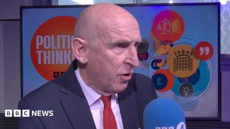 A headshot of Healey in the Political Thinking studio. He is bald and wears a navy suit with a white shirt and a red tie. Behind him is a Political Thinking backdrop and he is speaking into a blue BBC Radio 4 branded microphone.