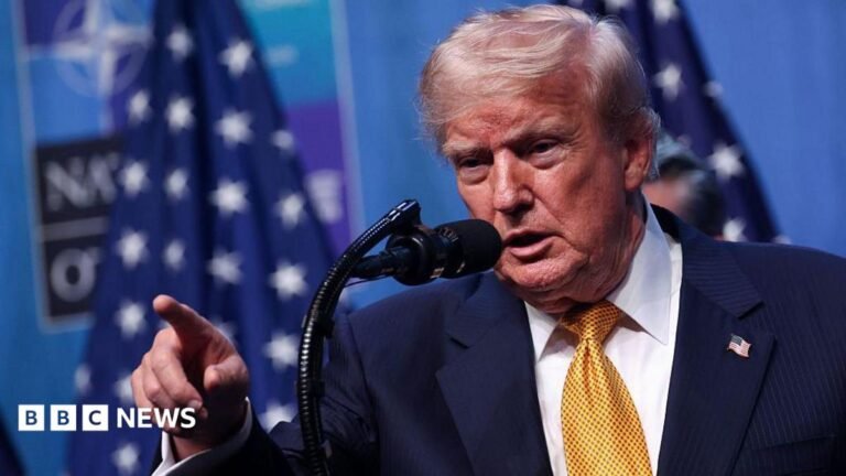 US President Donald Trump wearing a yellow tie, white shirt, blue suit and American flag pin. He stands at a microphone and is pointing, his mouth open.