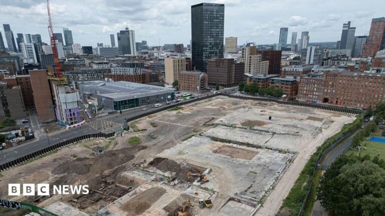 An aerial shot of two diggers in the middle of a large piece of empty land. There are buildings on the edges of the land, as well as an area of green space. In the background, there are clusters of high-rise towers.