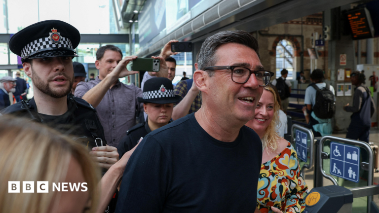 Who is Andy Burnham? Ex-Manchester mayor who wants to lead the country Andy Burnham flanked by two police officers. There are crowds of people taking photos behind him. He is wearing glasses and a dark shirt.