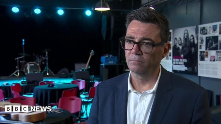 Andy Burnham. He has dark hair, wearing glasses, a navy blazer and white shirt.