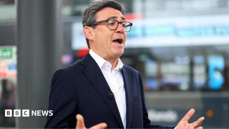 Andy Burnham says he’s not embarrassed about hoax message exchange Andy Burnham is wearing a dark-coloured jacket, white shirt and glasses. He has short black and grey hair. He is stood in front of two microphones and