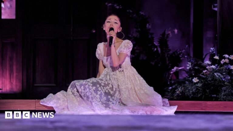 Ariana Grande sits on the stage, surrounded by flowers, as she sings on her Eternal Sunshine tour