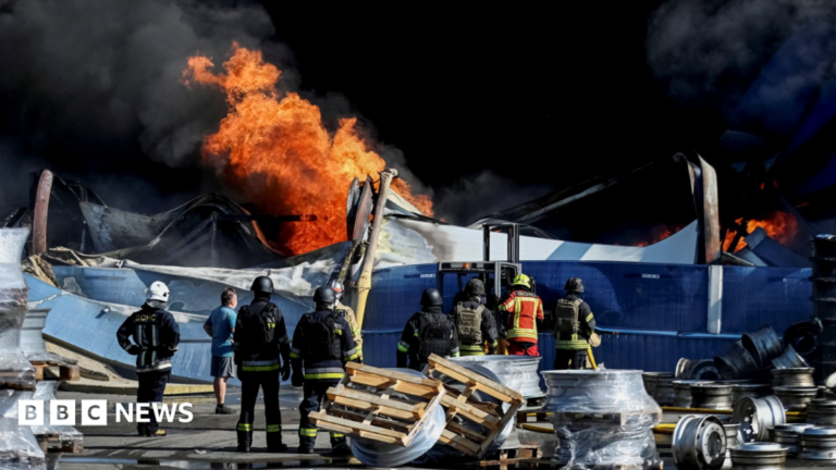 At least 37 dead and hundreds evacuated after strike on Kyiv warehouse Wide shot showing Ukrainian firefighters in black and yellow uniforms observing flames rising from the debris of what appears to be a warehouse following a drone strike.