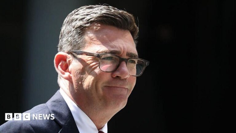 Andy Burnham is wearing glasses, a dark-coloured suit jacket, white shirt and a tie. He has a short, dark hair.