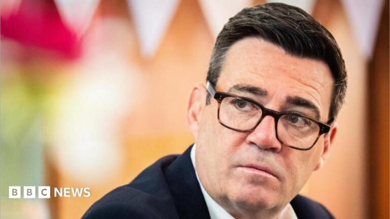Prime Minister Andy Burnham wearing a dark suit and glasses.