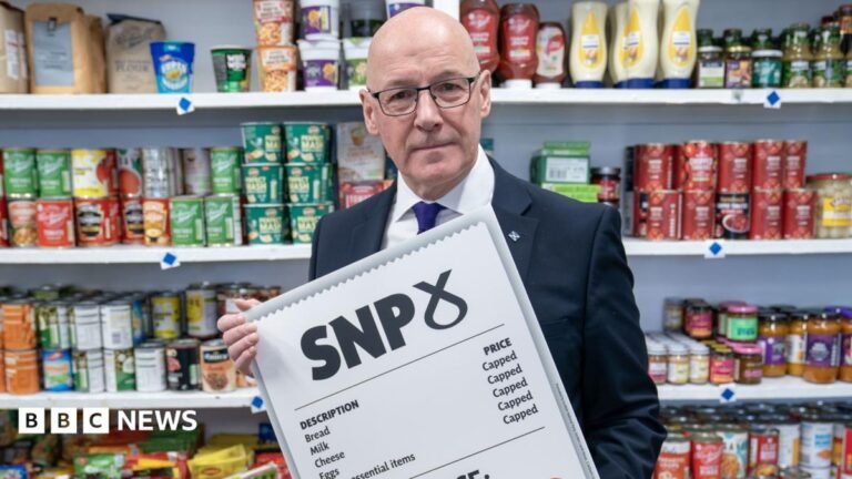 Business groups urge Swinney to scrap ‘ineffective’ food price cap plan A man smiles as he holds up a phone through which the US president is addressing the audience