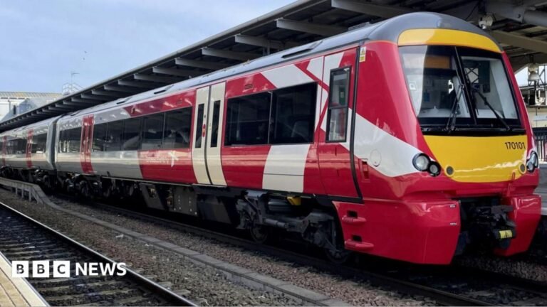 A red and white train stands at a train station. A platform can be seen in the background and another rail track in the foreground.
