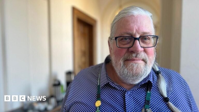 Devon councillor sanctioned in ‘sexualisation of children’ row Michael Fife Cook is wearing glasses and a blue and white shirt. He is wearing a lanyard and has grey hair in a ponytail and a grey beard and is standing in a corridor at County Hall offices.