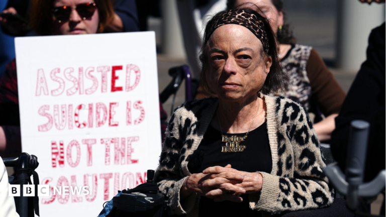 Actor Liz Carr, wearing a leopard print cardigan, black top and necklace. Next to her is a banner with the words 'Assisted suicide is not the solution'