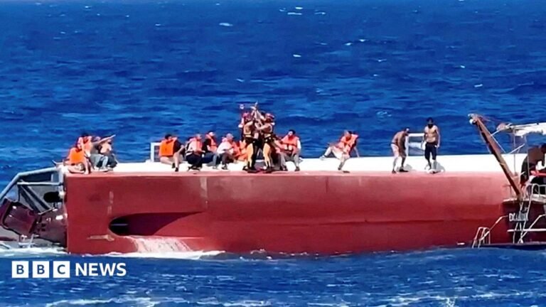Eight killed and others missing after ferry capsizes off northern Cyprus A close-up photograph shows a man and a woman - both with brown hair - posing together outdoors and looking toward the camera. The man wears a white hat. The backdrop is a city street with a building façade, outdoor seating and an orange scooter visible in the background.