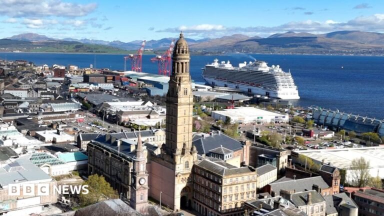 Greenock: The town whose council wants migrants to move in A coastal cityscape with a tall clock tower in the foreground, surrounded by historic and modern buildings. Behind it, a large white cruise ship is docked beside red industrial cranes. Beyond the harbour, green hills and mountains rise under a partly cloudy blue sky.
