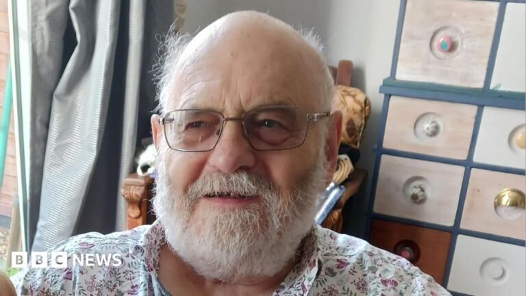 Ron Bailey, a man with a white beard and glasses, and a bald head. He is wearing a patterned open neck shirt and sitting next to a window in his home