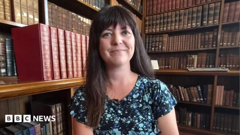 I was told I should not be at the National Eisteddfod, says Reform MS Helen Jenner is sat surrounded by books on shelves, with a large stack of red old-style books to the left of her head.